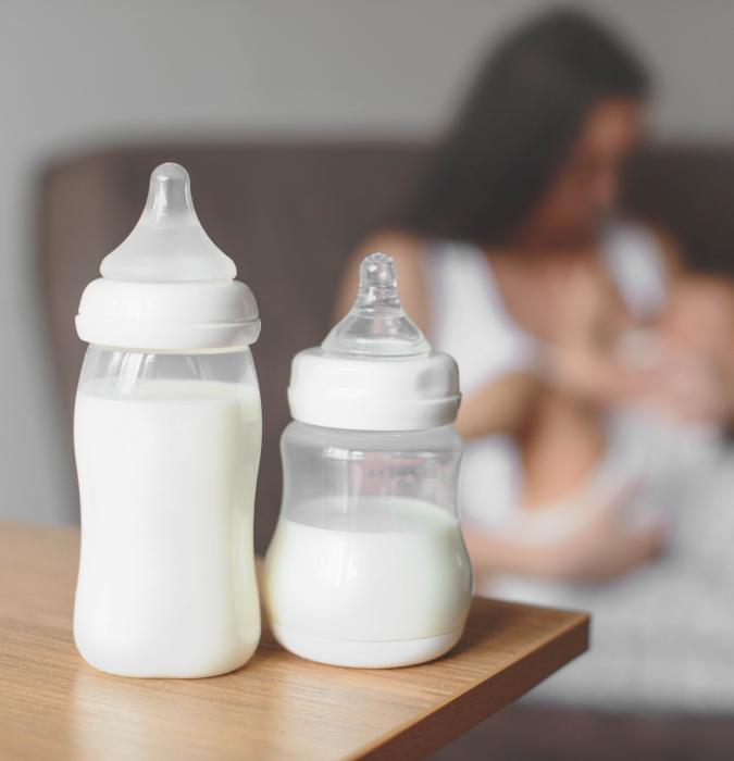Baby bottles full of milk sit in the foreground as a mother breastfeeds in the background. 